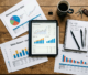 Tablet showing business performance charts surrounded by printed reports, a notebook with handwritten goals, pens, coffee mug, and keys